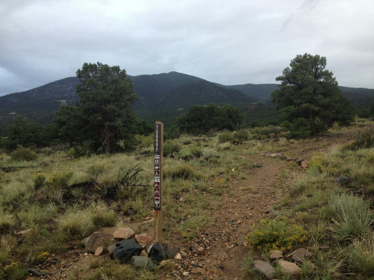 A dirt hiking trail leads through a grassy area with scattered rocks and shrubs, backed by green mountains under a cloudy sky. A wooden trail sign marks the path, indicating various recreational activities allowed in the area. Double Rainbow mountain bike trail.
