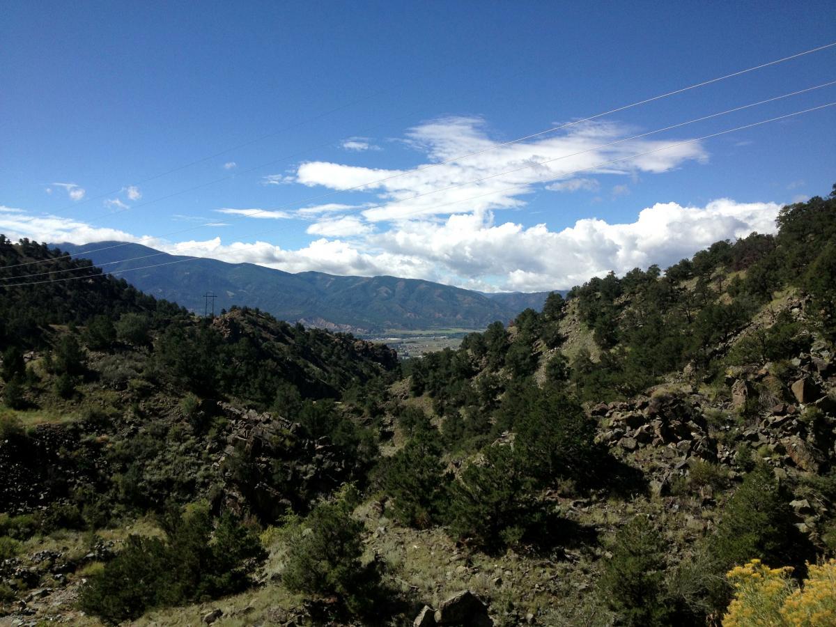A scenic view of a mountainous landscape with rolling hills covered in green vegetation and scattered rocks. The sky is bright blue with fluffy white clouds, and power lines are visible in the distance. The valley below shows a mix of nature and a hint of civilization. Cottonwood mountain bike trail.