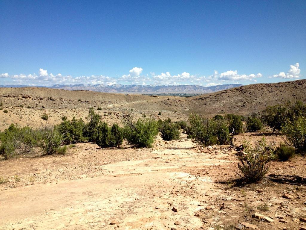 A panoramic view of a rocky landscape under a clear blue sky, featuring sparse vegetation and distant mountains. The terrain appears dry and rugged, with hills and valleys visible in the background. Mary's Loop / Horsethief Bench mountain bike trail.