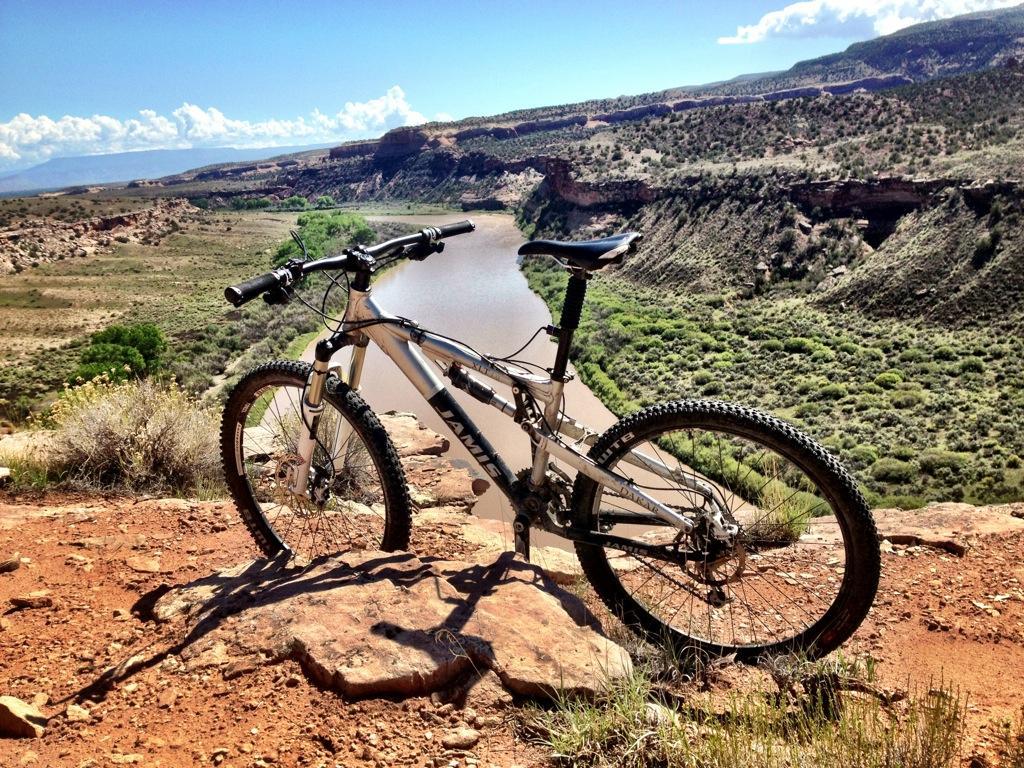 A mountain bike resting on a rocky ledge, overlooking a winding river surrounded by lush greenery and rugged terrain under a bright blue sky with scattered clouds. Mary's Loop / Horsethief Bench mountain bike trail.