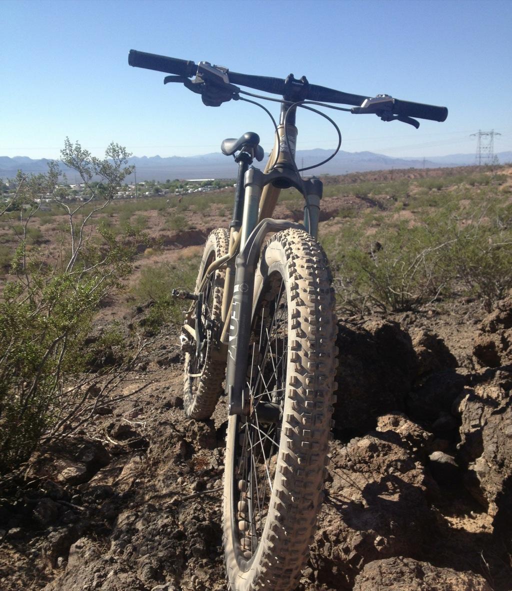 Surly Instigator: A mountain bike resting on rocky terrain, with dust-covered tires, set against a clear blue sky and distant mountains. Sparse vegetation surrounds the bike, suggesting a rugged outdoor environment.