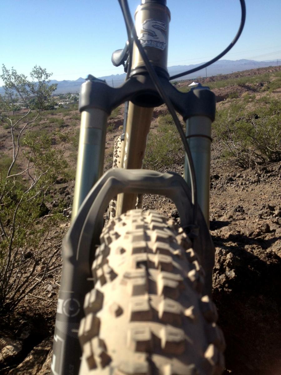 Surly Instigator: A close-up view of a mountain bike's front tire and suspension fork, positioned on a rocky trail with a mountainous landscape in the background under a clear blue sky. The tire shows signs of use with visible tread patterns, and sparse vegetation is seen surrounding the trail.