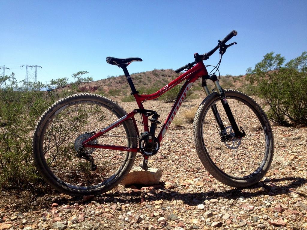 Niner Jet 9: A red mountain bike resting on a rocky terrain with sparse vegetation in a desert landscape, under a clear blue sky.