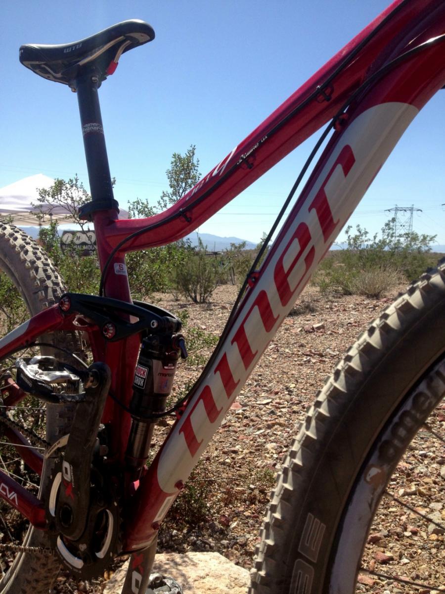 Niner Jet 9: A close-up view of a red mountain bike showing its frame, seat, and rear suspension. The bike is positioned outdoors with a clear blue sky and sparse vegetation in the background, indicating a rugged terrain suitable for biking.