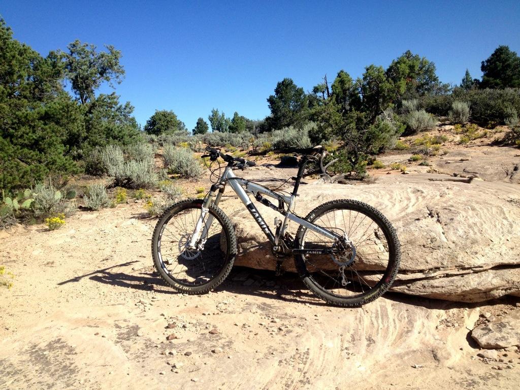 Alt text: A mountain bike resting on a rocky terrain with shrubs and trees in the background under a clear blue sky. Gooseberry Mesa mountain bike trail.