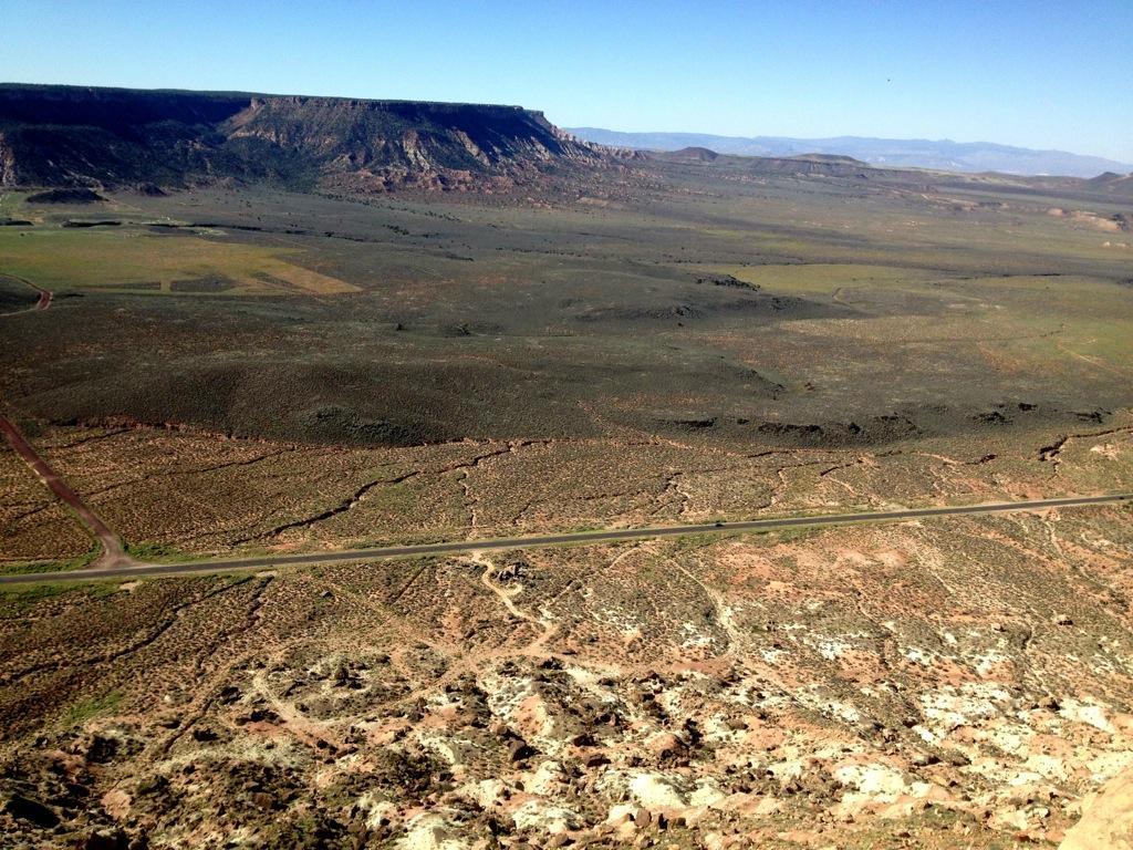A panoramic view of a vast desert landscape featuring rugged cliffs in the background, open fields in the foreground, and a winding road cutting through the terrain. The sky is clear, indicating bright weather, while the earthy tones of the ground contrast with the distant mountains. Gooseberry Mesa mountain bike trail.