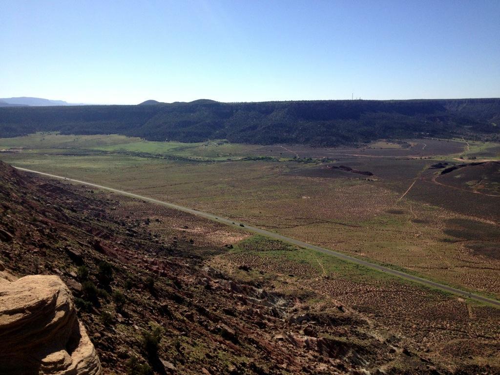 A panoramic view of a wide, open landscape featuring rolling hills and a distant mountain range under a clear blue sky. A winding road can be seen cutting through the grassy valley, surrounded by sparse vegetation and rocky terrain. The sunlight brightens the scene, highlighting the natural beauty of the area. Gooseberry Mesa mountain bike trail.