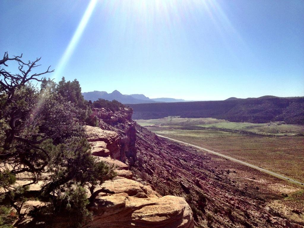 A scenic view from a rocky cliff, showcasing a vast, open landscape with distant mountains and valleys under a clear blue sky. Sunlight beams down, illuminating the greenery and earthy tones of the terrain below. Gooseberry Mesa mountain bike trail.