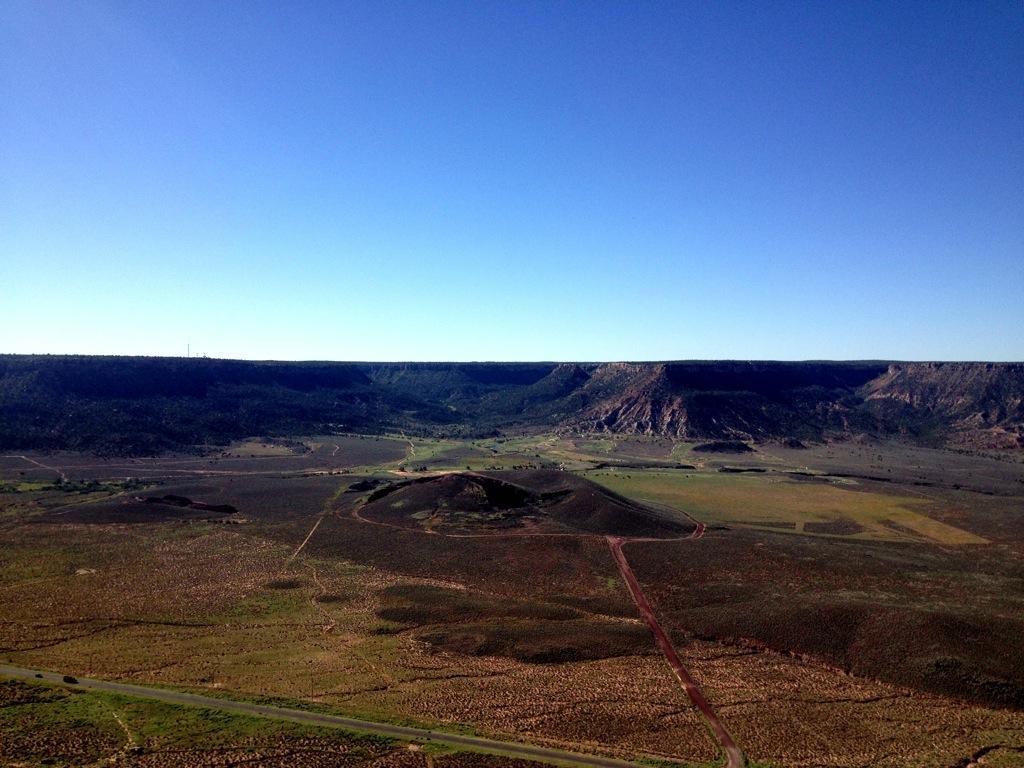 A panoramic view of a large, open valley surrounded by rugged hills and cliffs under a clear blue sky. The landscape features patches of green vegetation, dirt roads winding through the terrain, and a mix of grassy and rocky areas. Gooseberry Mesa mountain bike trail.