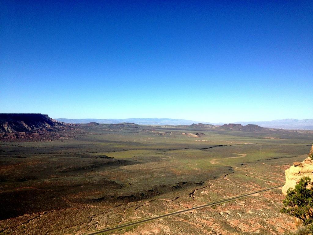 Wide view of a vast desert landscape under a clear blue sky, showcasing distant mesas and mountains, with a winding road cutting through the greenery and rocky terrain. Gooseberry Mesa mountain bike trail.