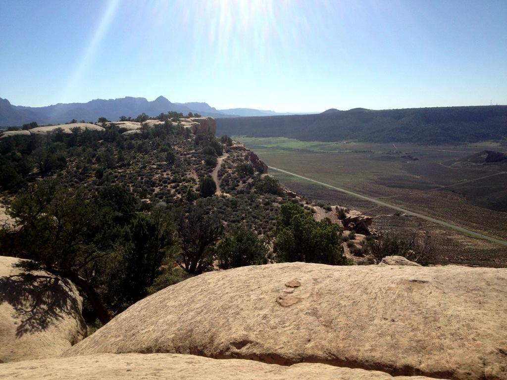 A panoramic view of a rugged landscape featuring rocky terrain, scattered vegetation, and distant mountains under a clear blue sky. Sunlight illuminates the scene, emphasizing the natural beauty of the area. A winding path can be seen cutting through the landscape below. Gooseberry Mesa mountain bike trail.