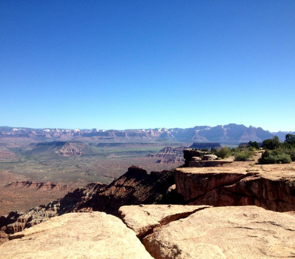 A panoramic view of a rugged desert landscape featuring layered rock formations and distant mountains under a clear blue sky. The foreground shows a rocky ledge with vegetation, while the expansive valley below is filled with varying terrain. Gooseberry Mesa mountain bike trail.