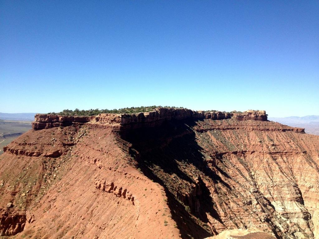 A rugged red rock formation under a clear blue sky, featuring steep cliffs and layered geological patterns, with a patch of greenery at the top. Gooseberry Mesa mountain bike trail.