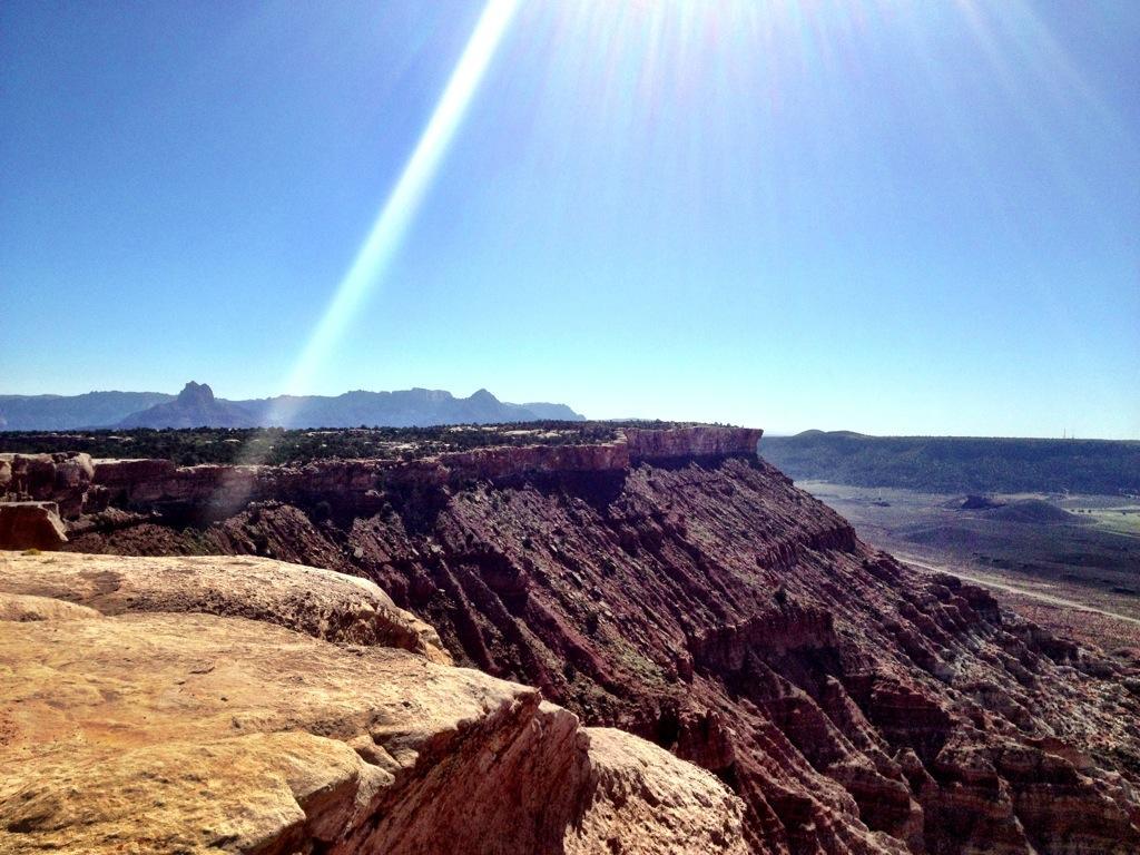A wide view of a rocky landscape under a clear blue sky, featuring rugged cliffs and distant mountains. The sun shines brightly, casting a glare and highlighting the textures of the rock formations. The scene conveys a sense of vastness and natural beauty. Gooseberry Mesa mountain bike trail.