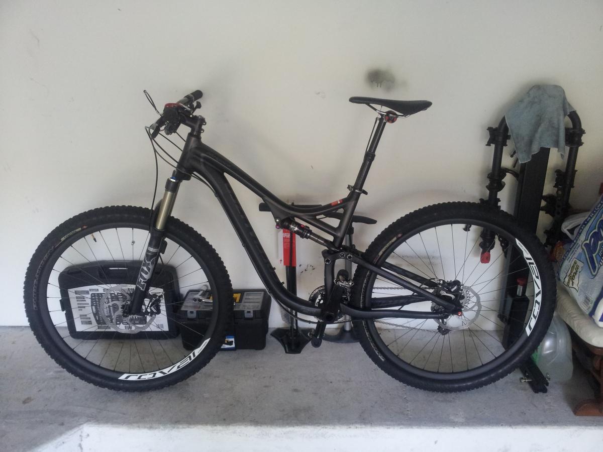 Specialized Stumpjumper FSR Comp 29: A black mountain bike leaning against a wall in a garage, featuring wide tires and a front suspension fork. In the background, there are tools and equipment, including a pump and various storage containers. The space is well-lit and organized.