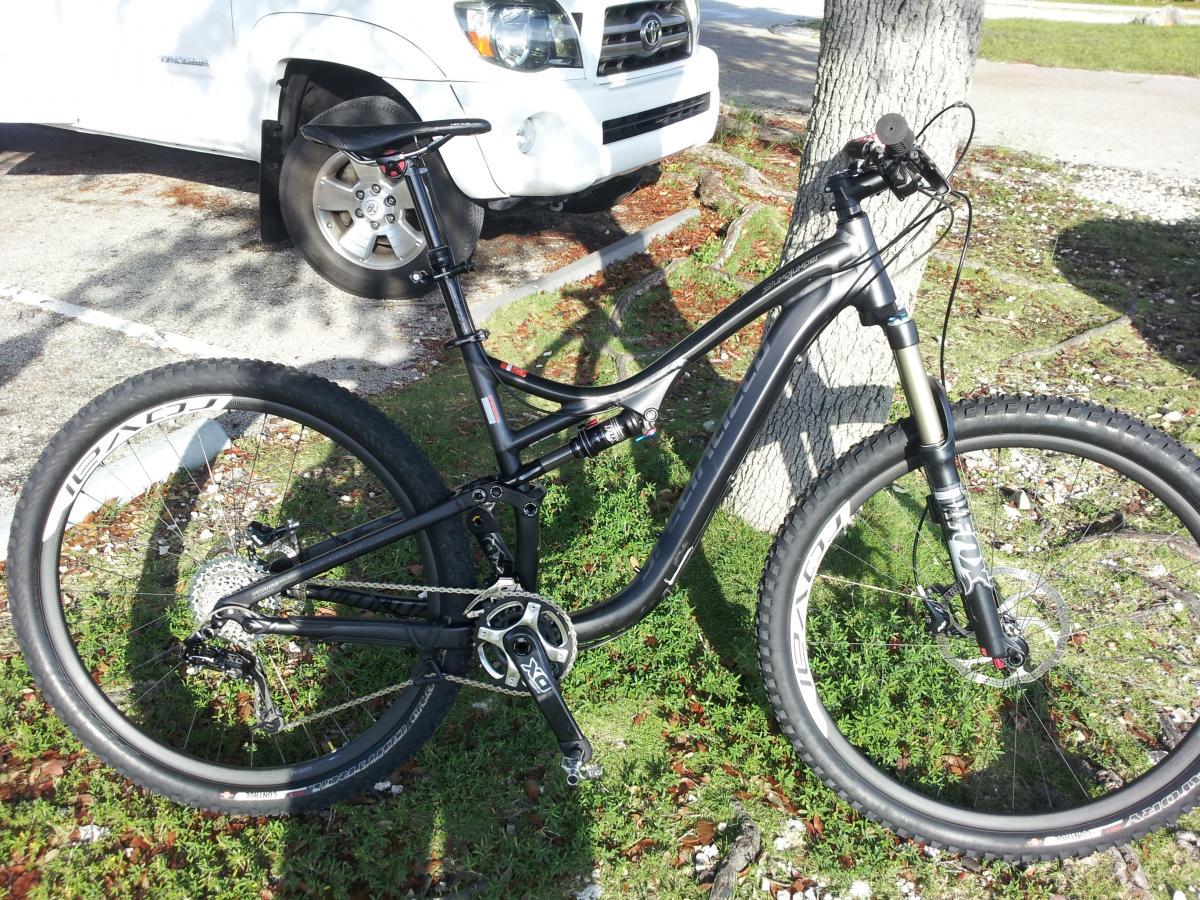 Specialized Stumpjumper FSR Comp 29: A black mountain bike resting on grass beside a tree, with a white vehicle parked in the background. The bike features prominent suspension forks, wide tires, and a modern design suited for off-road cycling.
