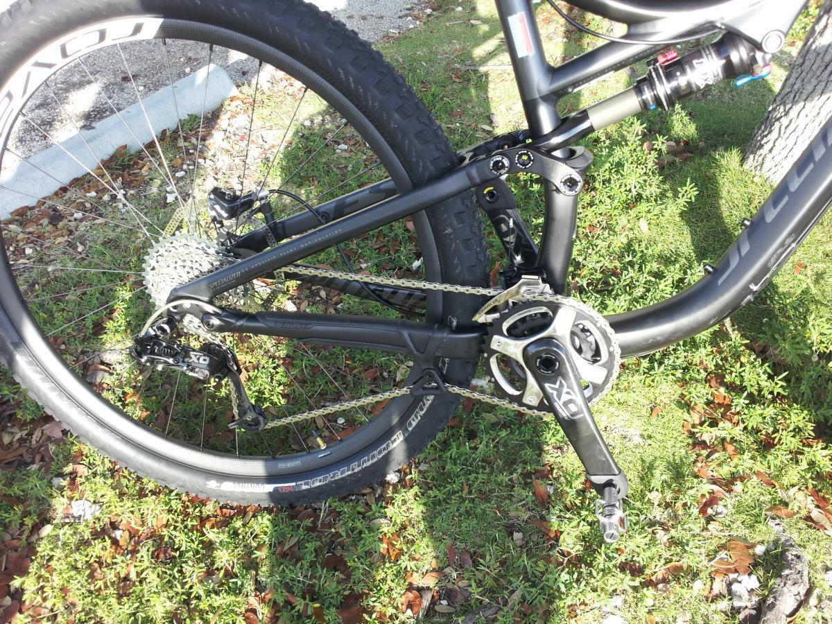 Specialized Stumpjumper FSR Comp 29: A close-up view of the rear section of a mountain bike, showing the chain, crankset, and rear derailleur. The bike features a black frame and specialized components, with a background of green grass and scattered leaves.