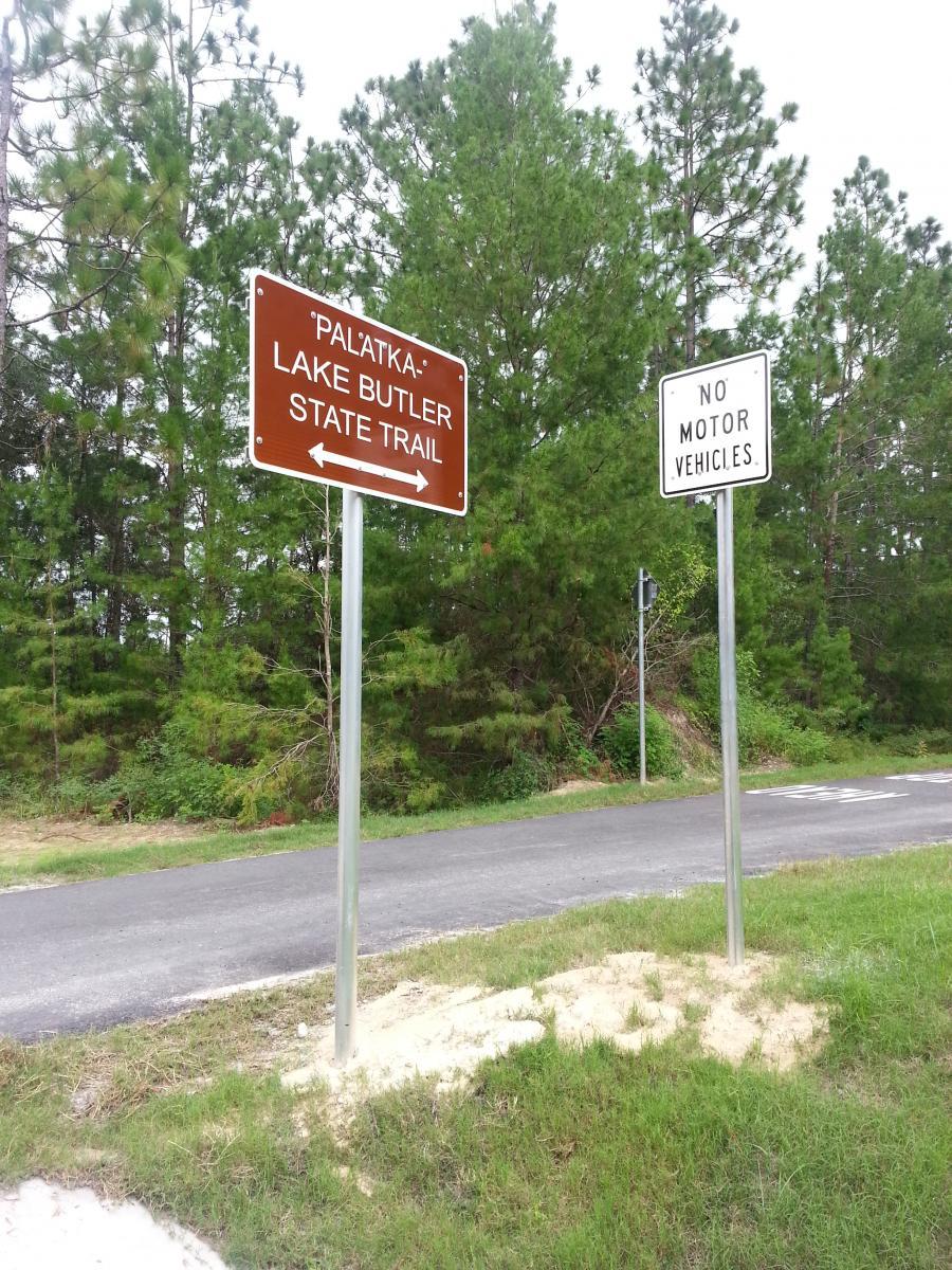 Two signs stand alongside a paved path in a forested area. One sign indicates the direction to the "Palatka Lake Butler State Trail" with arrows pointing left and right. The other sign states "No Motor Vehicles." Lush greenery surrounds the signs, highlighting a natural setting. Lake Butler to Plaka Trail mountain bike trail.