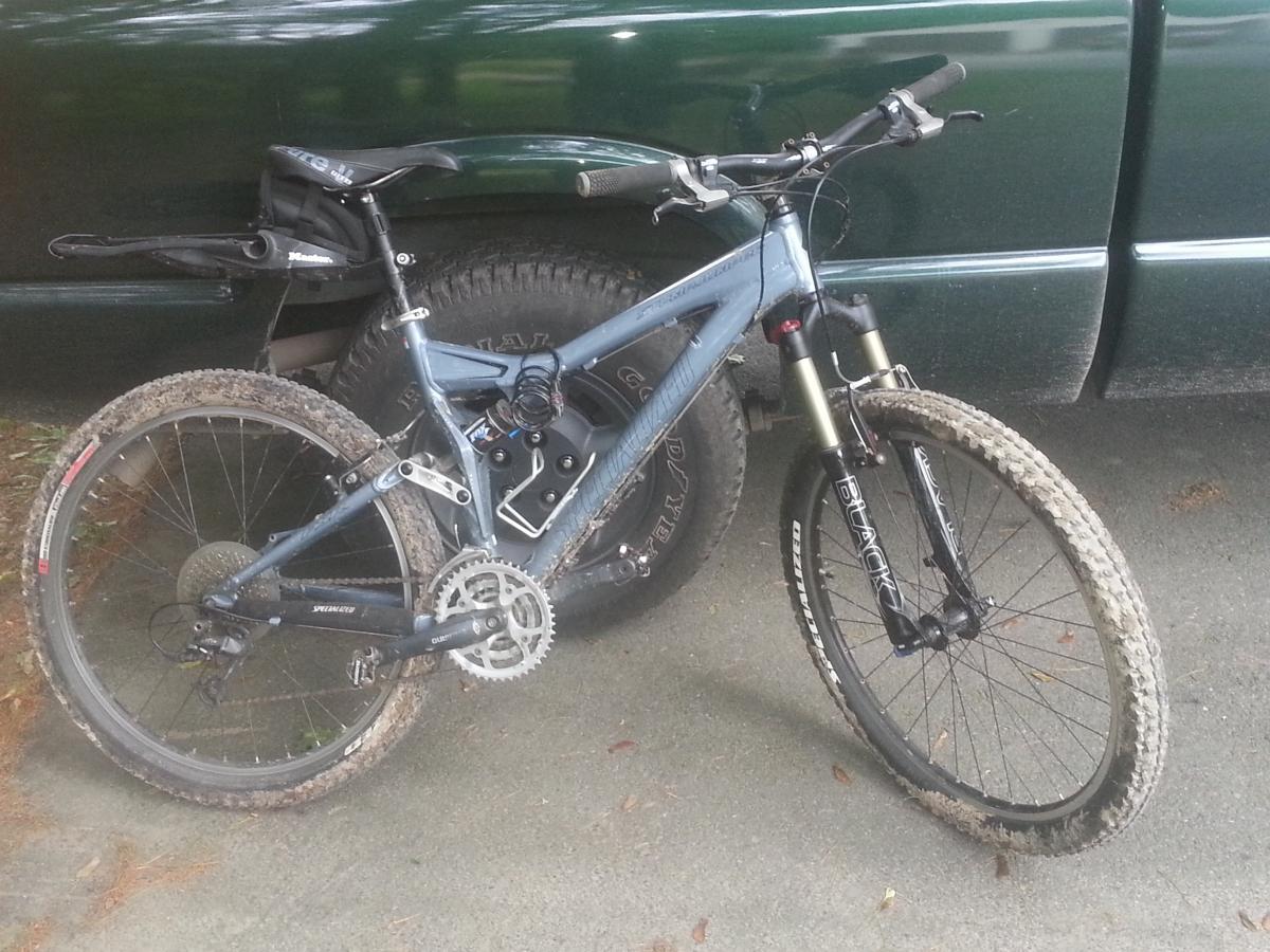 Specialized Stumpjumper FSR: A mountain bike with a blue frame, muddy tires, and a black seat, leaning against a green truck tire. The bike features front suspension and a gear system, indicating it is designed for off-road riding. The ground shows signs of dirt and leaves, reflecting an outdoor environment.