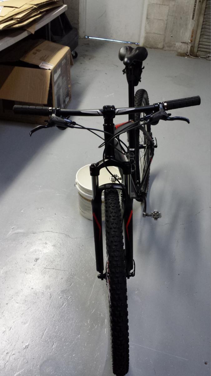 Specialized Hardrock Sport Disc 29er: A mountain bike viewed from the front, positioned on a smooth, light-colored floor. In the background, there are cardboard boxes and a closed door, suggesting an indoor storage space. A white bucket is partially visible under the bike. The bike features a black frame with red accents and knobby tires, indicating it's designed for off-road use.