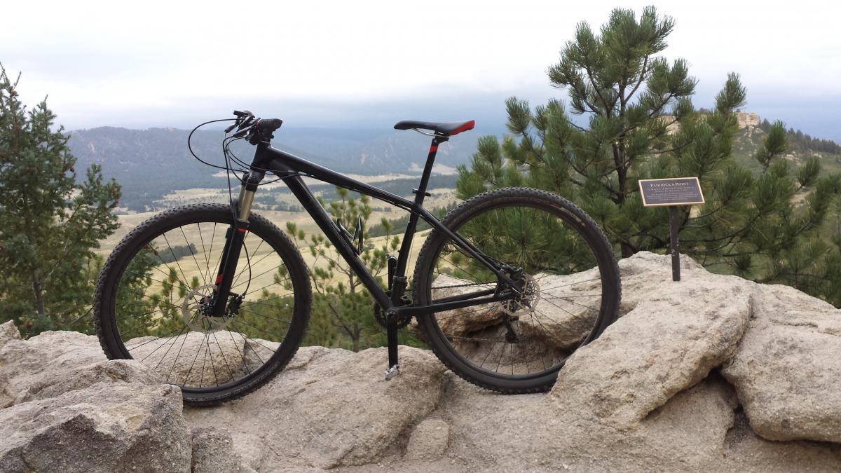 Specialized Carve Expert 29er: A mountain bike rests on a rocky outcrop, overlooking a scenic view of rolling hills and pine trees. In the background, there are distant mountains under a cloudy sky, and a sign positioned nearby indicates the location.