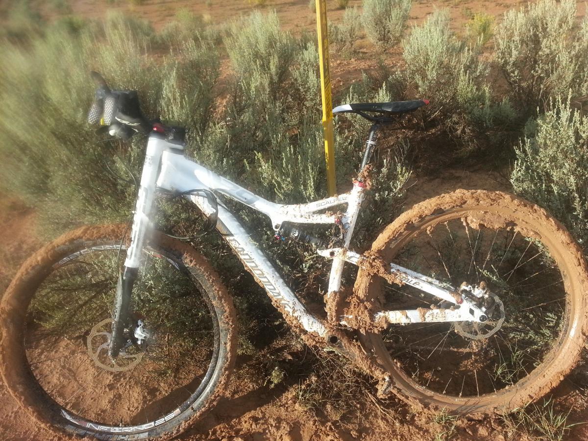 A muddy mountain bike leaning against a yellow post, surrounded by sparse greenery and dirt. The bike features a white frame, with its tires caked in mud, indicating it has been used on a challenging trail. Alien Run Trail mountain bike trail.