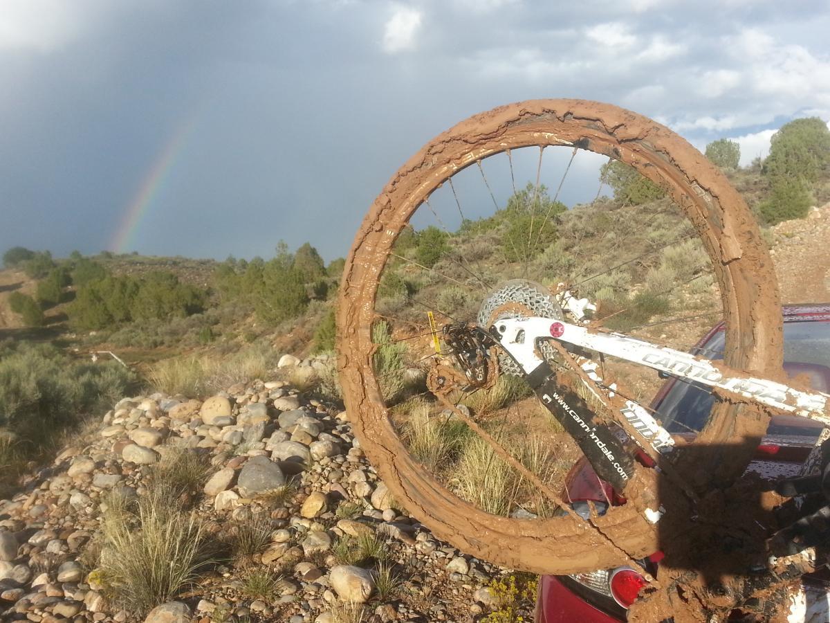 A muddy mountain bike wheel resting against a rocky terrain with a rainbow visible in the background, showcasing a scenic landscape featuring shrubs and trees. Alien Run Trail mountain bike trail.