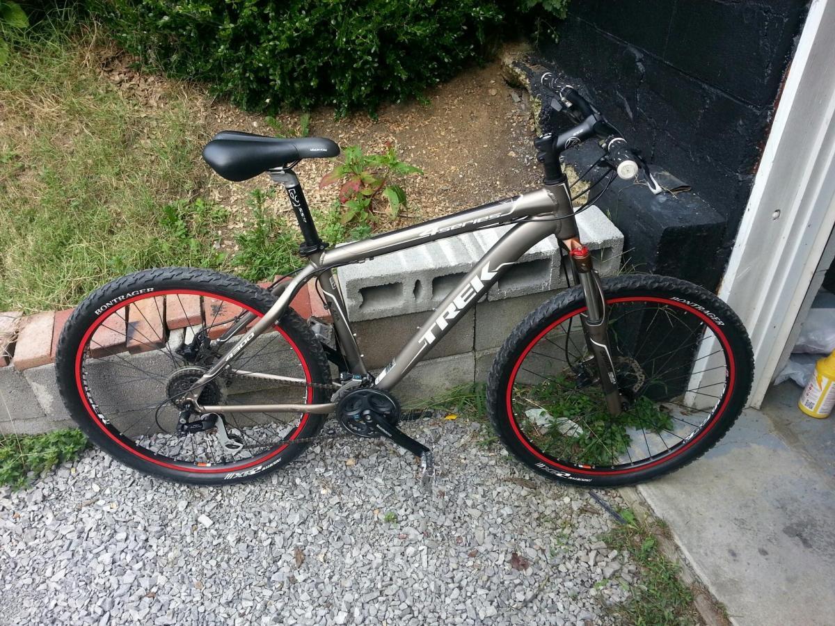 A Trek mountain bike parked on gravel, featuring a gray frame with red-accented wheels. The bike is positioned next to a concrete block, with grass and shrubs in the background.