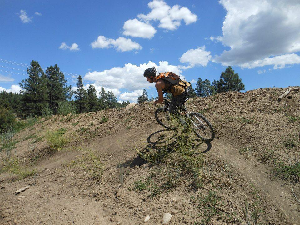 Specialized Rockhopper: A cyclist riding on a dirt path surrounded by trees under a blue sky with fluffy white clouds. The terrain is hilly, and the rider is leaning forward, wearing a helmet and an orange backpack.