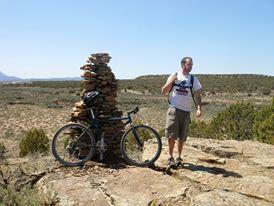 Specialized Rockhopper: A person standing next to a stack of rocks on a rocky landscape, with a bicycle nearby. The background features rolling hills and clear blue skies. The individual is wearing a t-shirt and shorts, and appears to be enjoying a sunny day outdoors.