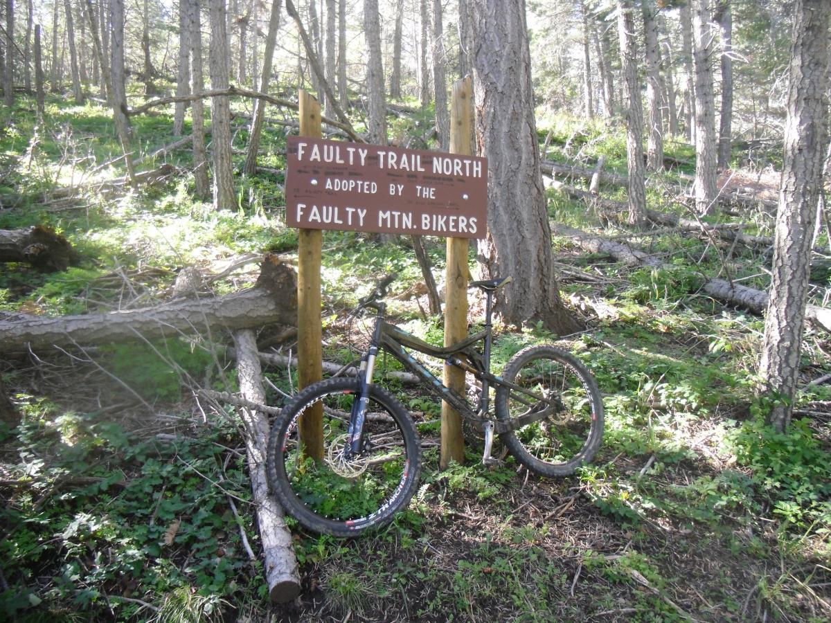 A mountain bike propped up against a sign that reads "Faulty Trail North - Adopted by the Faulty Mtn. Bikers," situated in a wooded area with trees and fallen branches in the background. Faulty Trail mountain bike trail.