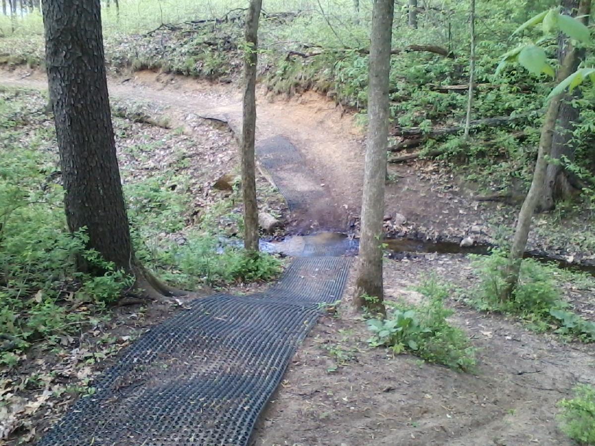 A narrow, textured path made of black matting leads through a wooded area, flanked by green plants and trees. The trail crosses over a small stream, with a dirt path curving off to the right in the background. The scene is lush and natural, indicative of a serene outdoor environment. Fort Custer Recreation Area mountain bike trail.
