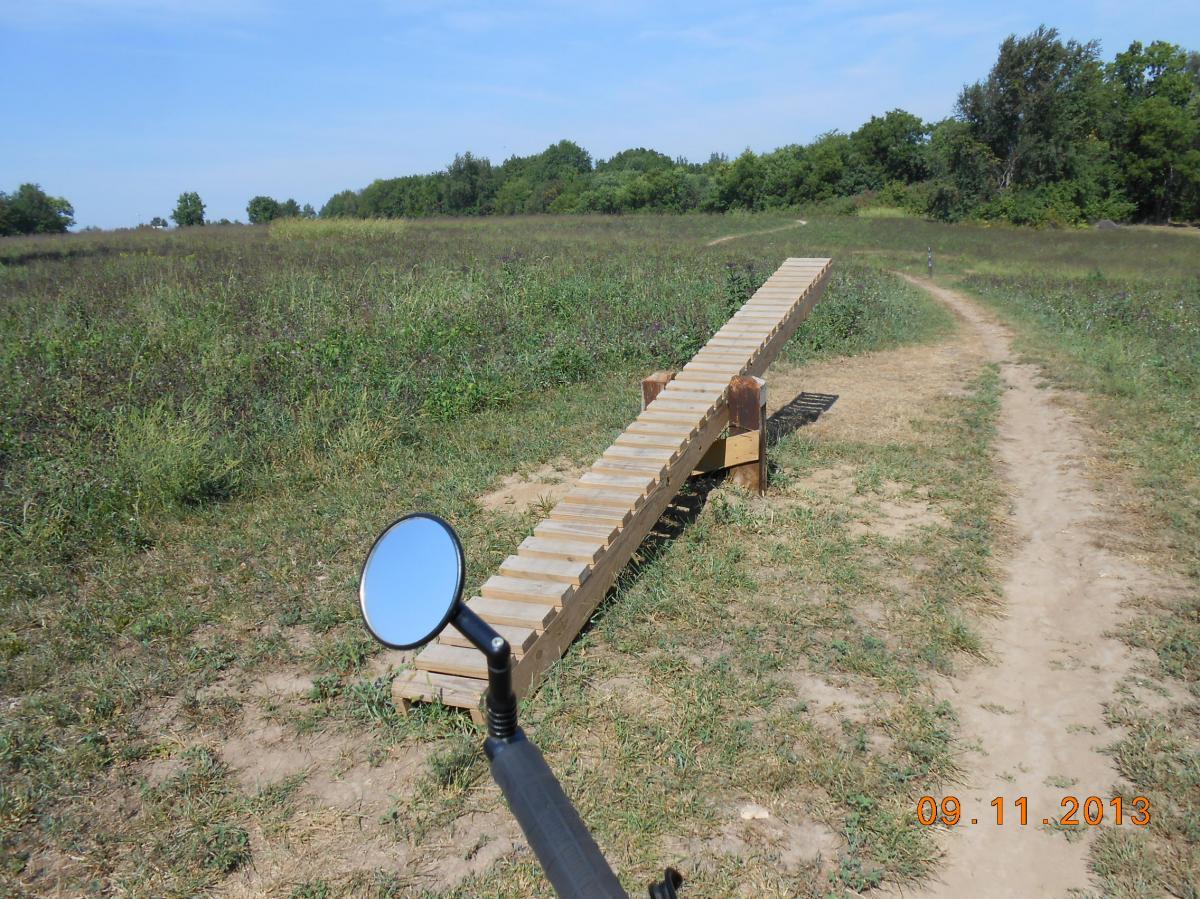 A wooden ramp leading into a grassy field, viewed from the side of a path. A bike mirror is visible in the foreground, reflecting the area. Blue sky and scattered trees are in the background. Date in the bottom corner shows September 11, 2013. Ruth Stockdale/walnut Woods mountain bike trail.