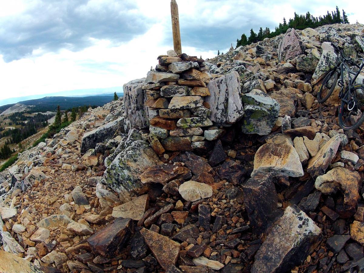 A rocky hillside with a stone cairn and a wooden marker at the summit, surrounded by sparse trees and distant mountains under a cloudy sky. A mountain bike is parked nearby on the uneven terrain. Medicine Bow Peak mountain bike trail.