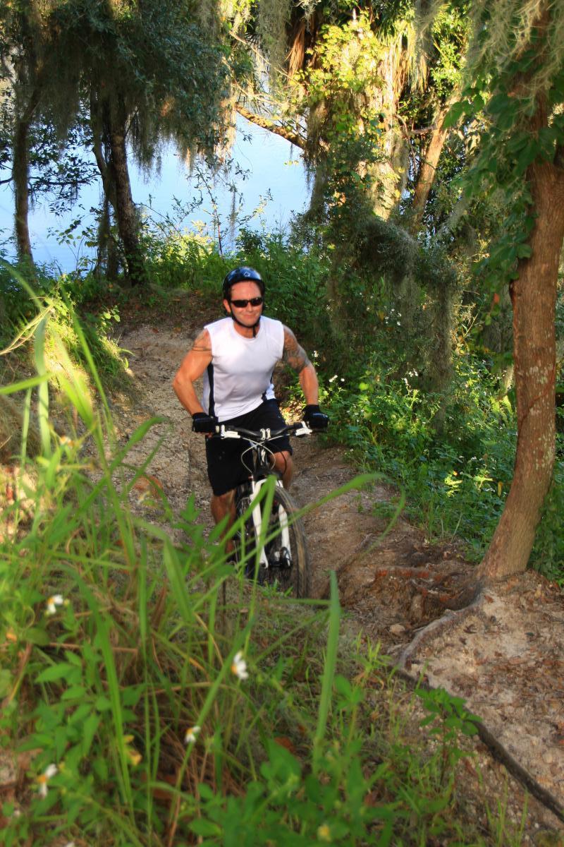 A cyclist riding uphill on a dirt trail surrounded by greenery and trees, with a river visible in the background. The rider is wearing a white sleeveless shirt, black shorts, and a helmet, smiling as they navigate the path. Loyce E. Harpe Park mountain bike trail.