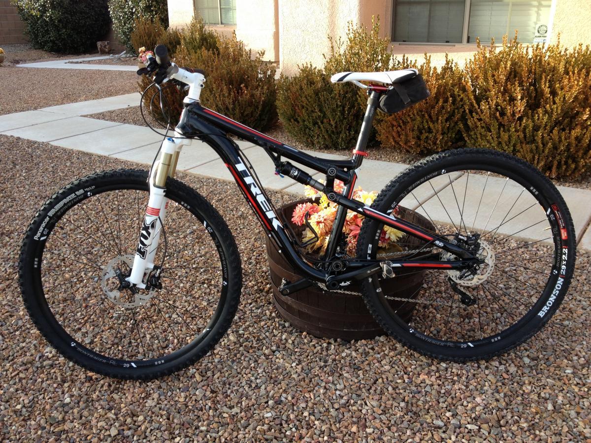 Trek Superfly 100 AL: Alt text: A black and red mountain bike leaning against a wooden planter, surrounded by gravel. The bike features wide tires and front suspension, with a small black bag attached to the seat. In the background, there are shrubs and a house with a window.