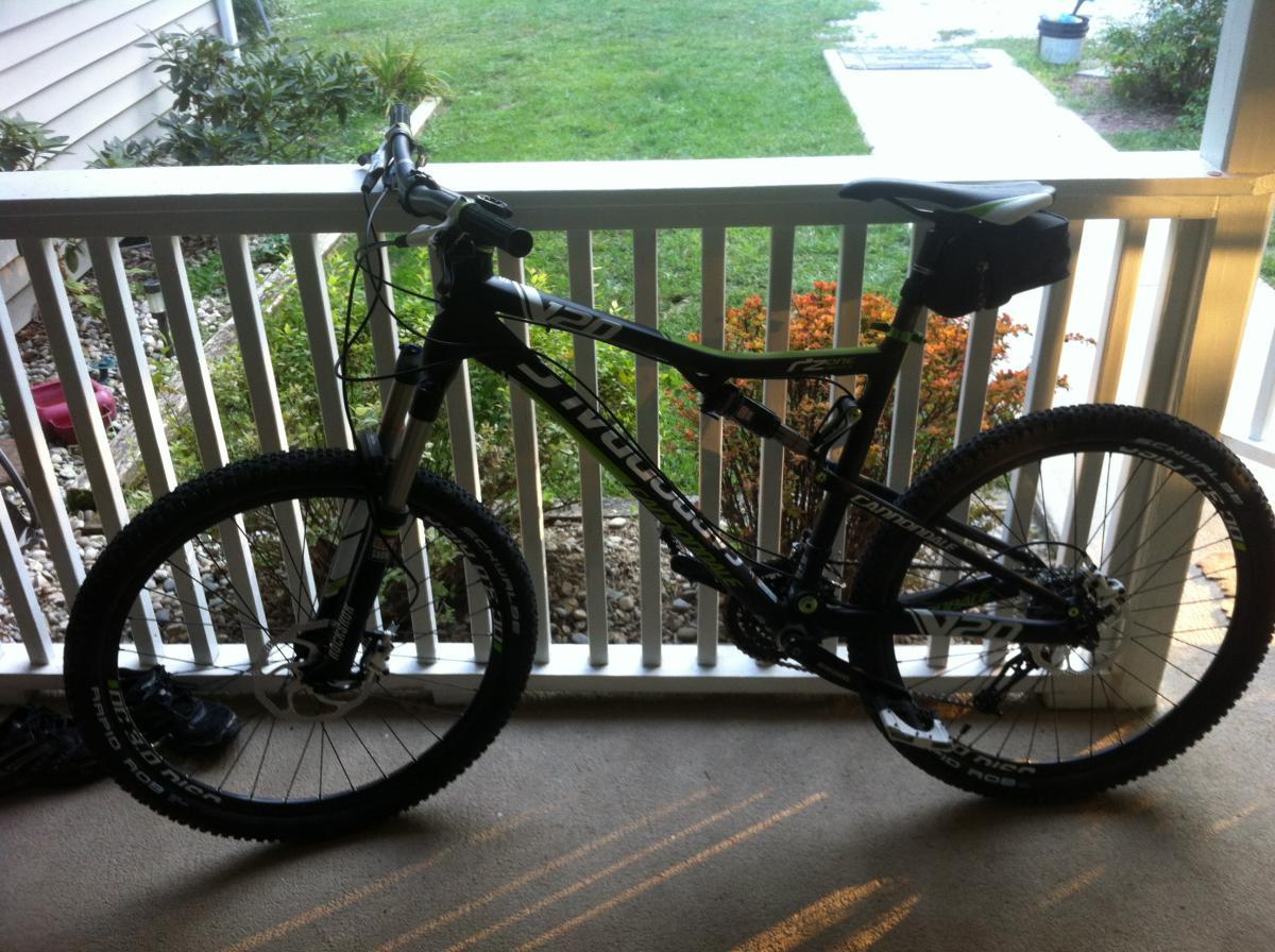 Cannondale RZ 120-1: Mountain bike parked on a porch, leaning against a railing. The bike features a black frame with green and white accents, wide tires, and is equipped with a small black saddlebag on the seat. In the background, there are plants and a patch of grass.