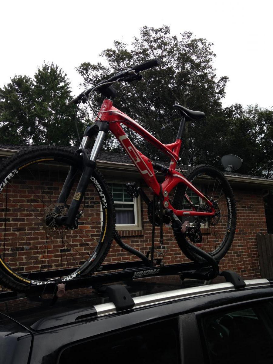 GT Force Carbon Pro: A red mountain bike secured on the roof rack of a black vehicle, set against a backdrop of a brick house and trees. The photo captures the bike from a low angle, showcasing its design and details.