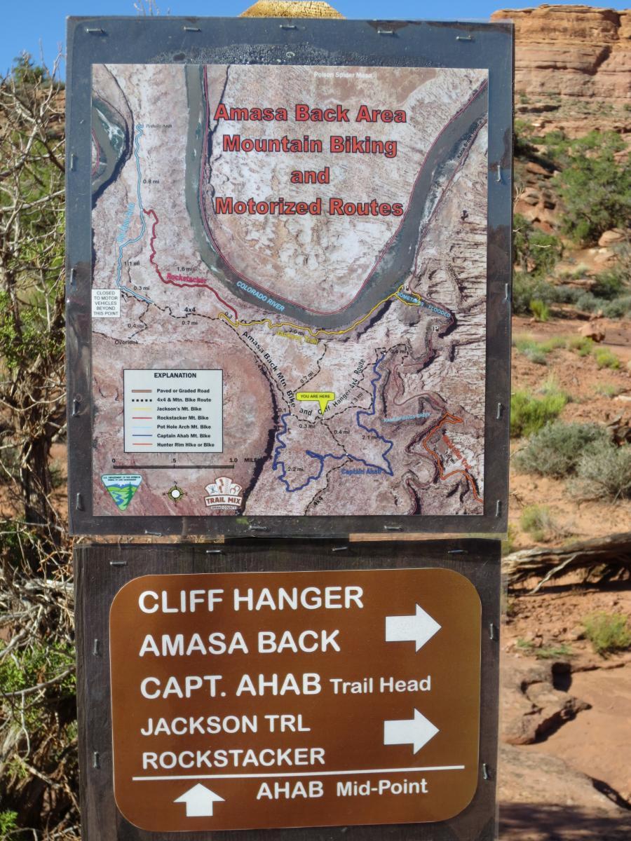 Signpost displaying a map of the Amasa Back Area, highlighting mountain biking and motorized routes. The map includes trail names and distances, with navigational arrows pointing towards various trailheads like Cliff Hanger, Capt. Ahab, Jackson Trail, and Rockstacker. The background features natural rock formations and greenery. Captain Ahab mountain bike trail.