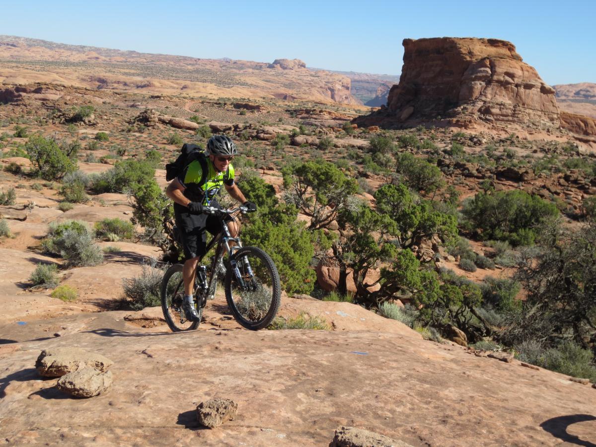 A mountain biker in a bright green shirt navigates a rocky trail in a desert landscape, surrounded by sparse vegetation and distant rock formations under a clear blue sky. Captain Ahab mountain bike trail.