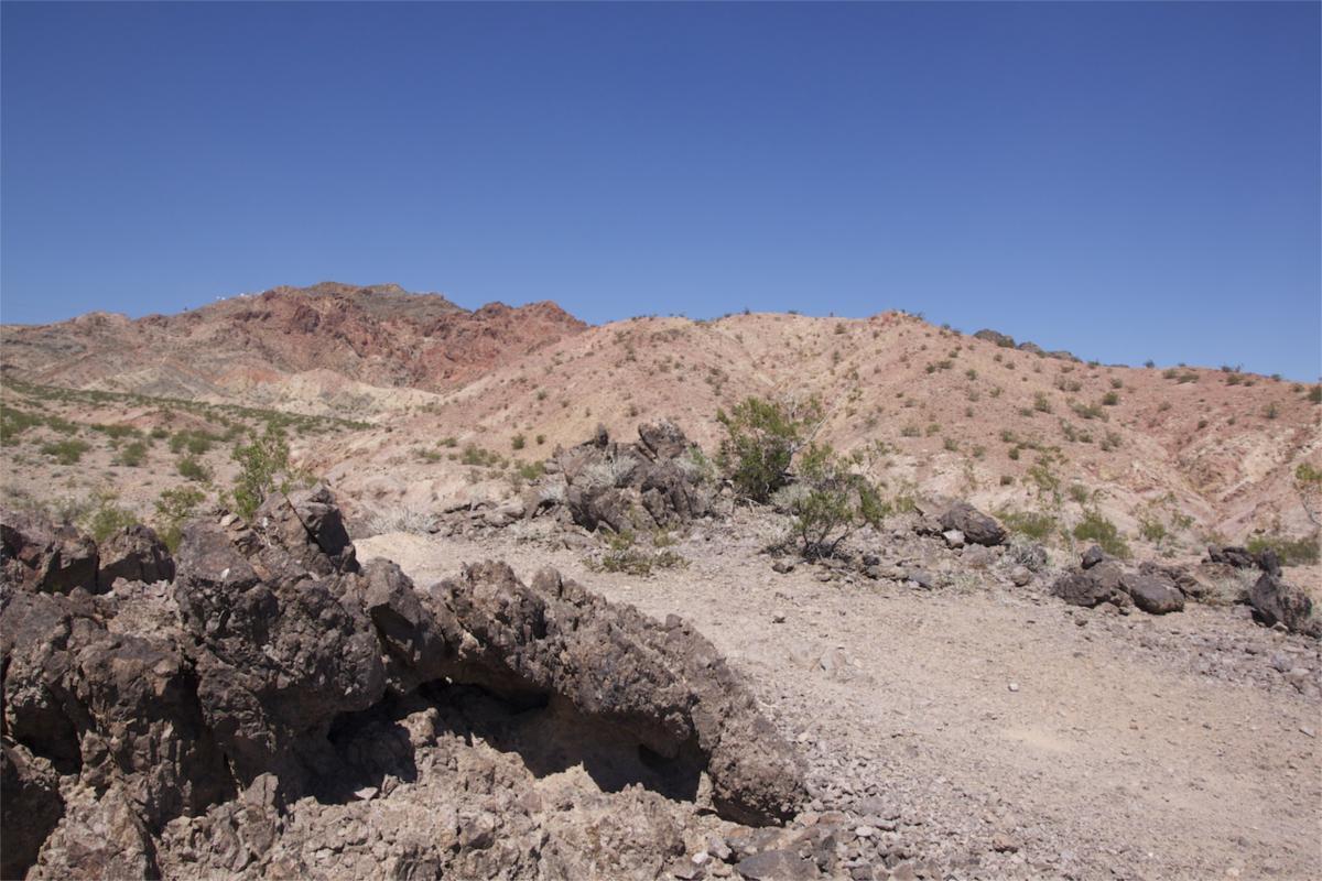 A dry, rocky landscape featuring rugged terrain and rolling hills in various shades of red and tan under a clear blue sky. Sparse vegetation is visible among the rocks, highlighting the arid environment. Bootleg Canyon mountain bike trail.
