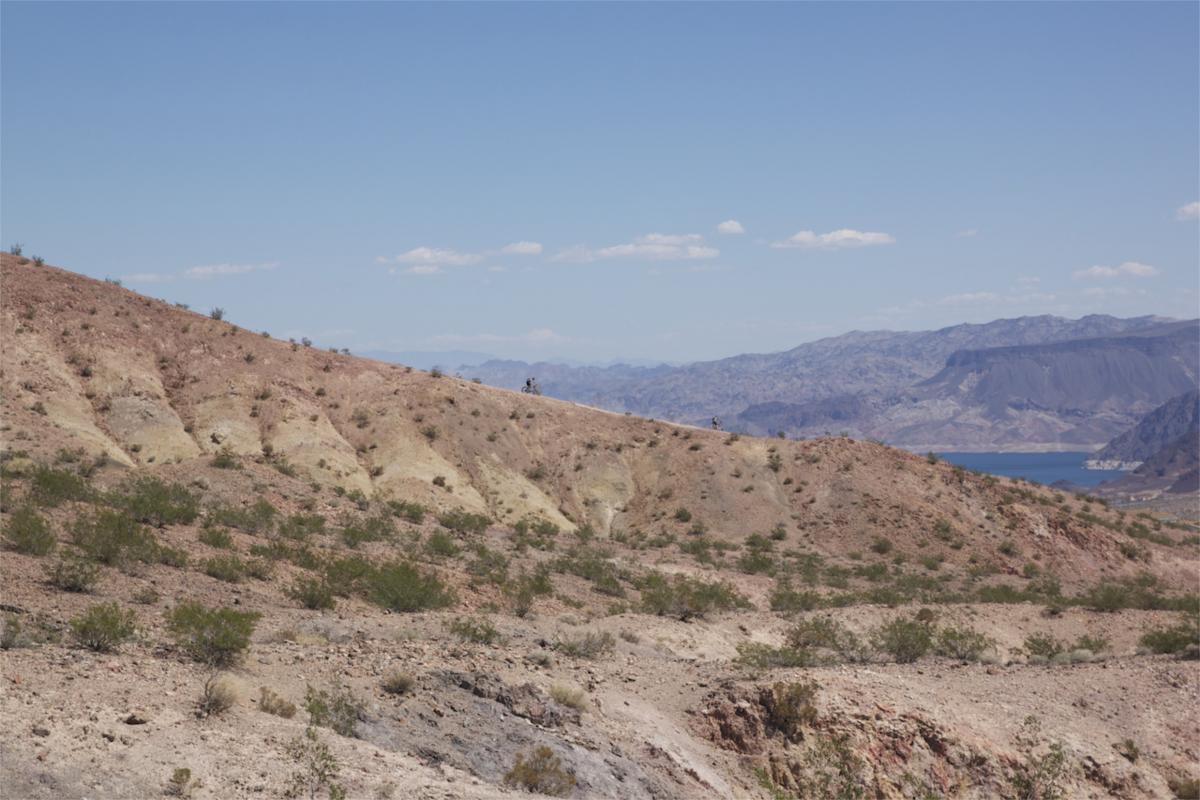 A panoramic view of a dry, hilly terrain featuring muted shades of brown and beige, dotted with sparse vegetation. In the background, a blue lake is visible surrounded by rugged mountains under a clear sky with a few scattered clouds. Two figures can be seen in the distance atop a hill, highlighting the vastness of the landscape. Bootleg Canyon mountain bike trail.