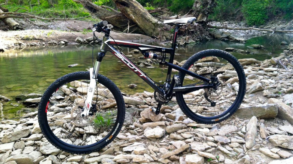 Scott Spark 60: A mountain bike resting on rocky terrain beside a calm stream, surrounded by lush greenery and fallen trees. The bike features a black and white frame with thick tires, indicating it is designed for off-road cycling.
