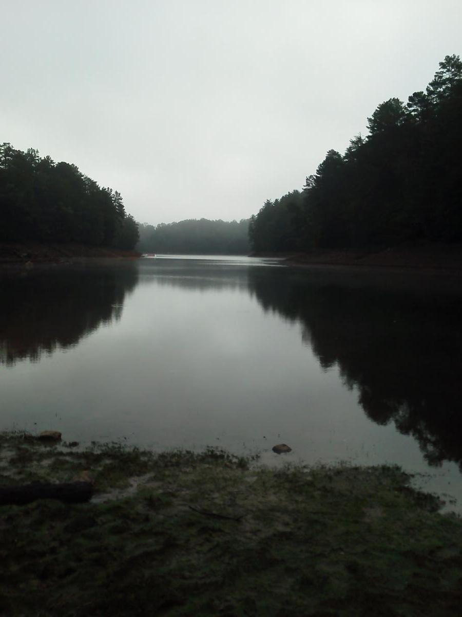 A calm, reflective lake surrounded by trees under a cloudy sky, with a sandy shoreline in the foreground. The water is still, mirroring the muted colors of the landscape and creating a serene atmosphere. Green Mountain mountain bike trail.