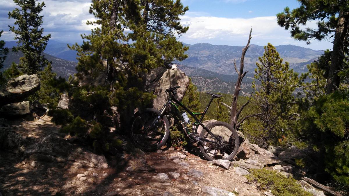 A mountain bike rests against a rock in a scenic outdoor setting, surrounded by pine trees and a mountainous landscape under a partly cloudy sky. Bergen Peak mountain bike trail.