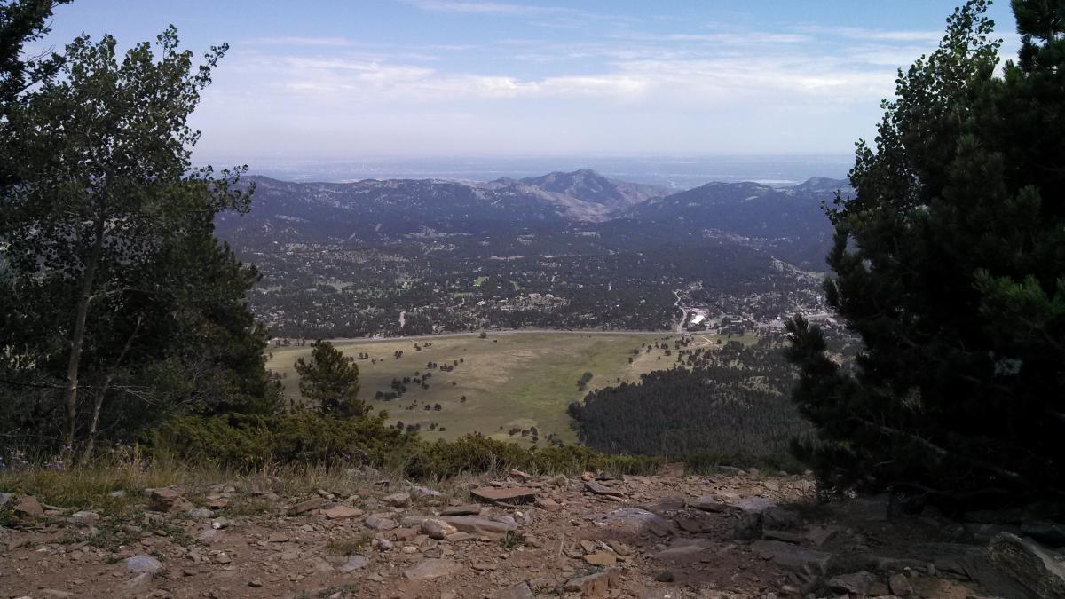 A scenic view from a mountain overlook, featuring lush green valleys and rocky terrain below, with distant mountains and a cloudy sky in the background. Bergen Peak mountain bike trail.