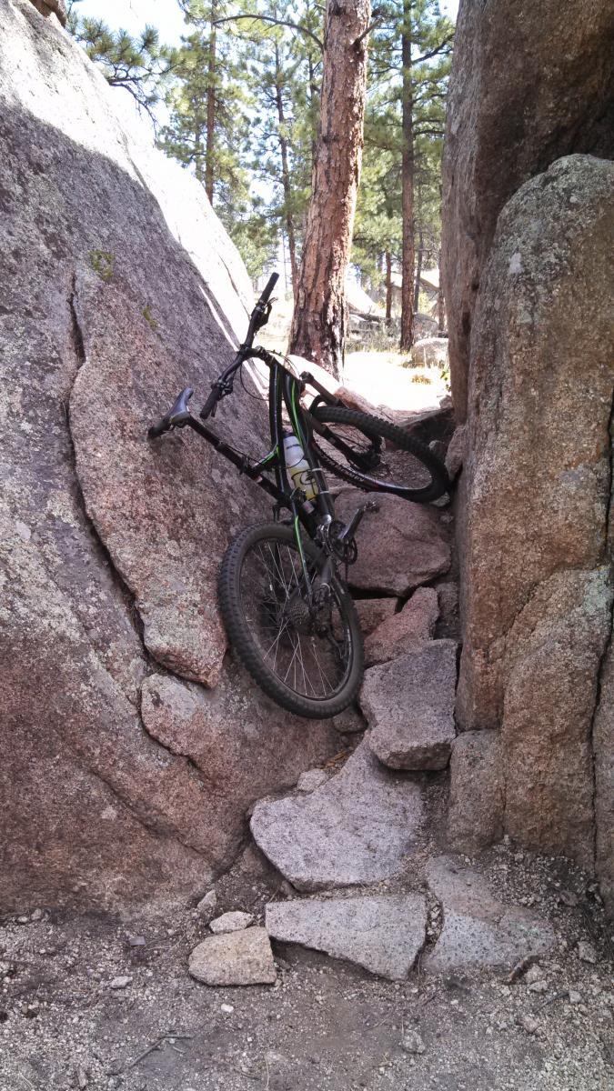 A mountain bike wedged between two large rock formations on a rugged trail, surrounded by pine trees. The area features rocky terrain with scattered boulders and a clear blue sky above. Blackjack / Raspberry Ridge mountain bike trail.