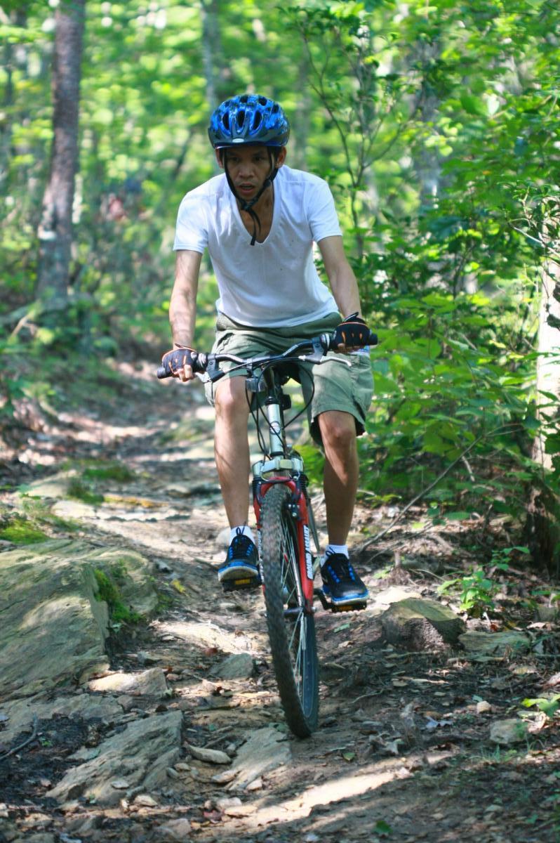 A person riding a mountain bike on a rocky trail surrounded by lush greenery. The cyclist is wearing a blue helmet and a white t-shirt, with shorts and athletic shoes. Sunlight filters through the trees, highlighting the rugged path and natural scenery. Coldwater Mountain mountain bike trail.