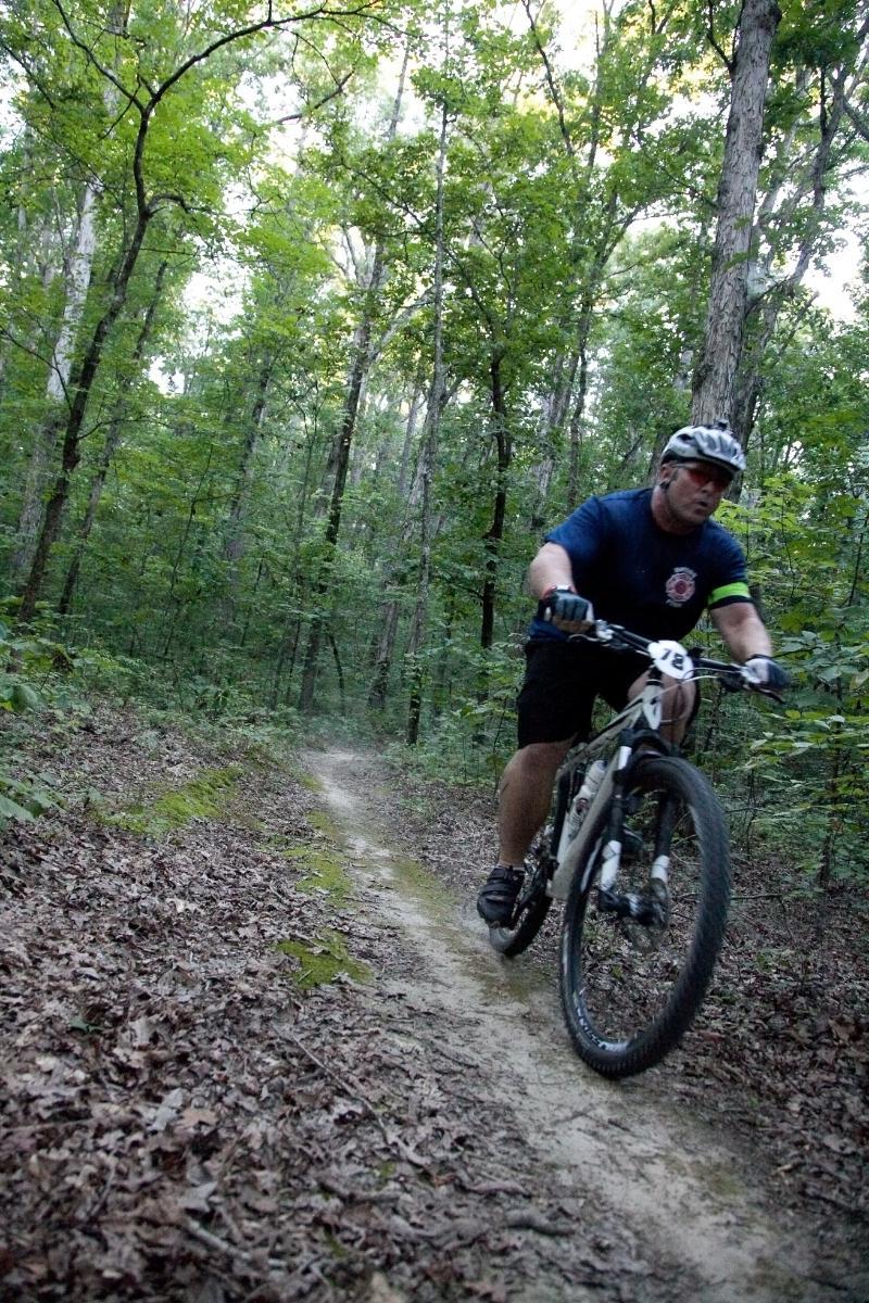 Specialized Carve Expert 29er: A mountain biker riding on a narrow dirt trail through a lush green forest, with trees and foliage surrounding the path. The cyclist is leaning forward on the bike, wearing a helmet and a blue shirt, as they navigate the terrain.