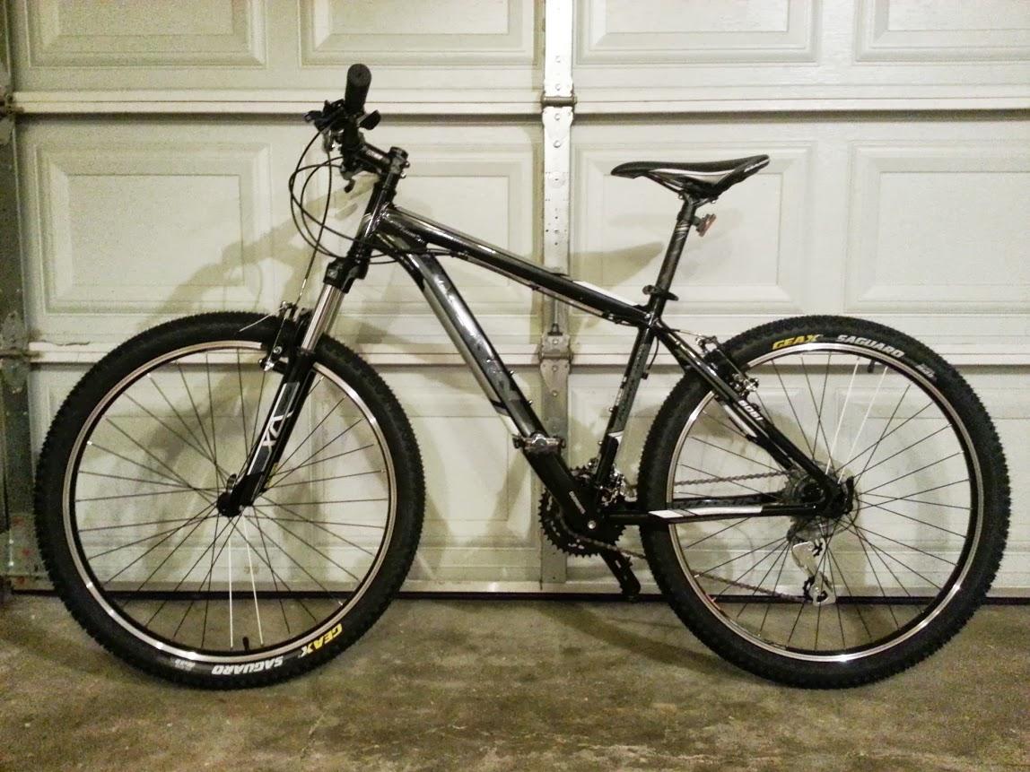 Marin Pioneer Trail: A black and silver mountain bike is leaning against a garage door. The bike features wide tires with pronounced treads, front suspension, and a sleek frame design. The setting is an indoor garage with a concrete floor and closed garage door in the background.
